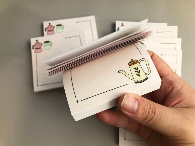 Puede incluir: Una pila de peque&ntilde;os blocs de notas cuadrados con una ilustraci&oacute;n dibujada a mano de una tetera. La tetera es de color crema con un asa y un pico marrones, y hojas verdes. Otros blocs de notas tienen ilustraciones de cafeteras y tazas.