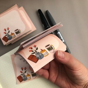 May include: Pink sticky notes with a floral and teacup illustration. The notes feature a soft pink border and the words "You are a gift" on one. A hand holds a stack of the notes, with pens in the background.