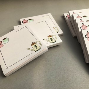 Puede incluir: Blocs de notas cuadrados blancos con tem&aacute;tica de caf&eacute;. Cada bloc de notas presenta un borde negro e ilustraciones de una cafetera rosa, una taza verde y una tetera amarilla. Los blocs de notas est&aacute;n apilados sobre una superficie gris.