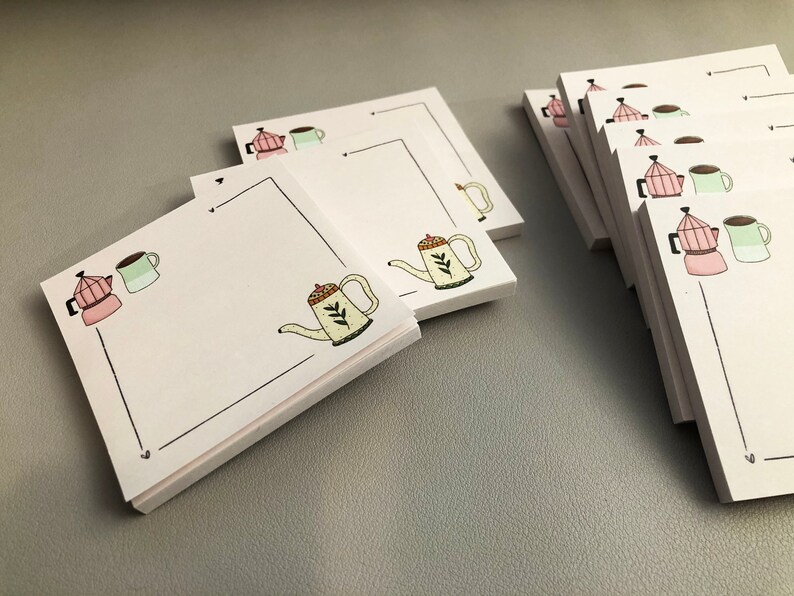 Puede incluir: Blocs de notas cuadrados blancos con tem&aacute;tica de caf&eacute;. Cada bloc de notas presenta un borde negro e ilustraciones de una cafetera rosa, una taza verde y una tetera amarilla. Los blocs de notas est&aacute;n apilados sobre una superficie gris.
