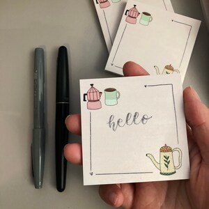 Puede incluir: Un bol&iacute;grafo gris y un bol&iacute;grafo negro est&aacute;n junto a una pila de blocs de notas blancos. Los blocs de notas tienen un tema de caf&eacute; con una cafetera rosa, una taza verde y una tetera amarilla. La palabra "hello" est&aacute; escrita en el bloc de notas.