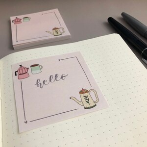 Puede incluir: Notas adhesivas rosas con un dise&ntilde;o dibujado a mano que presenta una cafetera, una taza y una tetera. La palabra "hello" est&aacute; escrita en cursiva. Una pila de notas similares y bol&iacute;grafos est&aacute;n en el fondo.