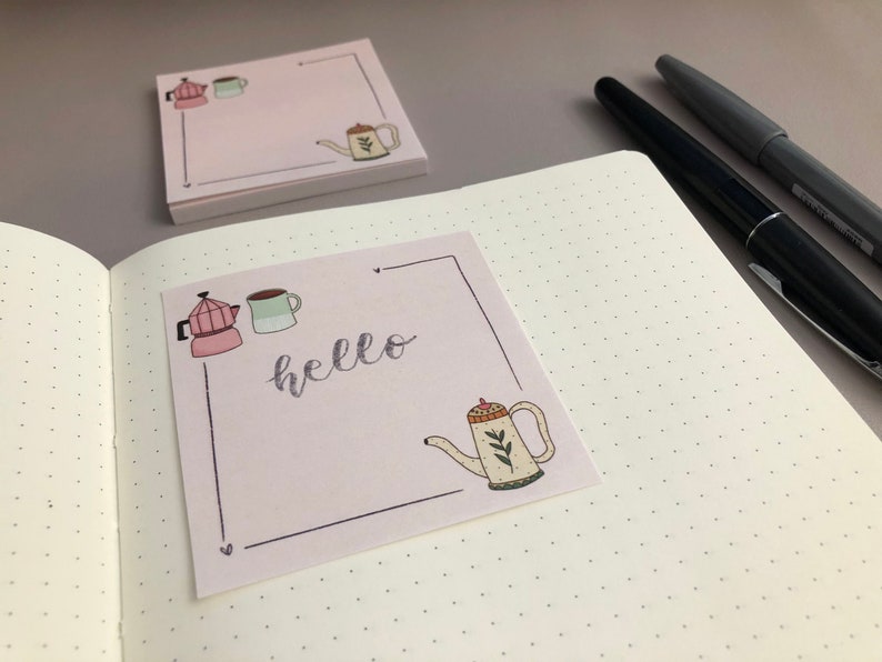 Puede incluir: Notas adhesivas rosas con un dise&ntilde;o dibujado a mano que presenta una cafetera, una taza y una tetera. La palabra "hello" est&aacute; escrita en cursiva. Una pila de notas similares y bol&iacute;grafos est&aacute;n en el fondo.