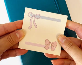 Peut inclure: Un petit post-it crème avec deux illustrations de nœuds roses et des rayures horizontales grises et roses. La note est tenue entre deux mains, avec un cahier bleu canard en arrière-plan.