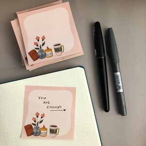 May include: Pink square sticky notes with a floral and coffee cup illustration, and the text "You are enough". A black pen and a gray marker are next to the notes. A blue notebook is also in the image.