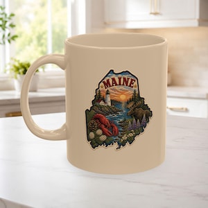 Pode incluir: Uma caneca de cerâmica bege com um gráfico detalhado do estado do Maine. O design inclui um farol, um pôr do sol sobre a água, um alce e uma lagosta vermelha, com a palavra MAINE impressa em letras vermelhas em negrito no topo.