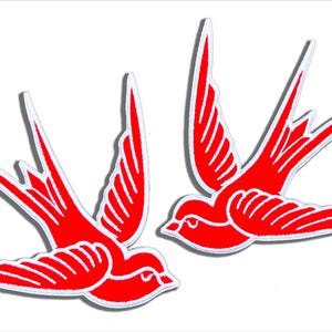 May include: Two embroidered swallow patches featuring a vibrant red color with white stitched outlines and detailing. Each bird is depicted in a flying position with wings spread and long, pointed tail feathers, creating a symmetrical pair of bird motifs.