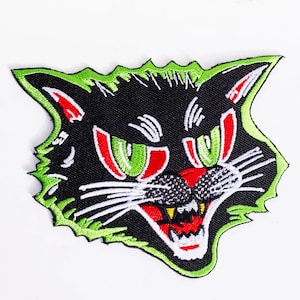 Könnte beinhalten: Ein schwarzes Katzen Gesicht mit grünen und roten Augen, einer weißen Nase und einem weißen Mund mit scharfen Zähnen. Das Katzen Gesicht ist grün umrandet und hat einen gezackten Rand.