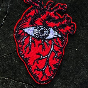 May include: Red embroidered heart with a black outline and a single eye in the center. The eye is purple and white with a black pupil.