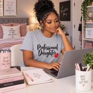 May include: A heather gray t-shirt with the black text Professional Daughter and a small red heart graphic. The shirt features a crew neckline and short sleeves, worn while typing on a laptop at a desk with a notebook, pens, and books.
