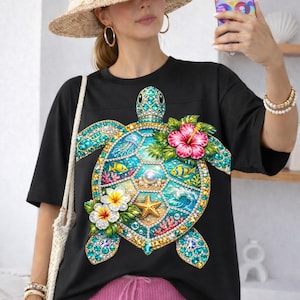Puede incluir: Camiseta negra con un diseño colorido de tortuga marina, adornada con gemas, flores y elementos oceánicos. La camiseta se combina con pantalones cortos rosas y un sombrero de paja.