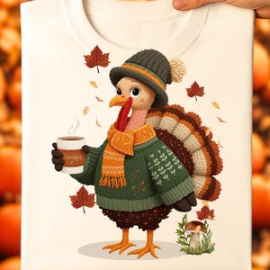 Könnte beinhalten: Weißes T-Shirt mit einer Cartoon-Truthahn, der einen grünen Pullover, einen orangefarbenen Schal und einen Hut trägt und eine Kaffeetasse hält. Der Truthahn hat einen braunen und weißen Federschwanz und ist von Herbstblättern umgeben. Auf der Tasse steht "Harvest".