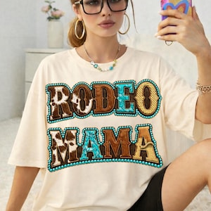 Może przedstawiać: Kremowy t-shirt z napisem "RODEO MAMA" w stylu western. Tekst ma brązowe i turkusowe akcenty, ze wzorem skóry krowy w literach. Koszulka jest w rozmiarze oversize i ma krótkie rękawy.