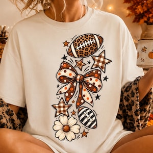 Puede incluir: Una camiseta color crema con un estampado gráfico de un balón de fútbol con estampado de leopardo, un lazo grande de lunares, una margarita y varias estrellas en tonos marrón, naranja y blanco, dispuestos verticalmente en la parte frontal.