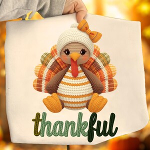May include: A white canvas tote bag featuring a crocheted turkey wearing a white hat with an orange bow. The turkey has orange, brown, and yellow feathers. The word "thankful" is written in green and black script at the bottom.
