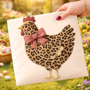Könnte beinhalten: Ein cremefarbenes quadratisches Kissen mit einem dekorativen Hühnerdesign. Das Huhn hat ein Leopardenmuster, einen roten Kamm, einen gelben Schnabel und eine rosa Schleife. Das Kissen wird vor einem verschwommenen Hintergrund im Freien gehalten.