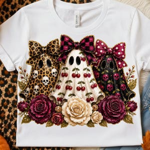 Op de afbeelding: Een wit T-shirt met korte mouwen en een afbeelding van drie geesten met grote strikken. De linkergeest heeft een luipaardprint met schedels, de middelste een kersenpatroon en de rechter een zwarte schedelprint. Onderaan staan donkerrode en crèmekleurige rozen.