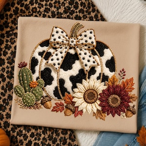 May include: A beige t-shirt featuring an embroidered design of a pumpkin with a black and white cow print pattern, topped with a polka dot bow. The pumpkin is surrounded by a green cactus, sunflowers in cream and maroon, and brown acorns.