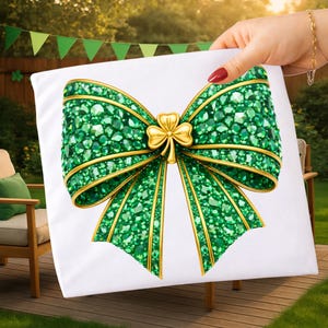 Op de afbeelding: Een wit kussen met een groot, sierlijk strikdesign. De strik is voornamelijk groen, met een gouden rand en een gouden klaver in het midden. De strik is bedekt met kleine, glinsterende groene edelstenen. Het kussen wordt voor een groen gazon gehouden.