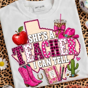 May include: A white t-shirt featuring a graphic of a Texas-shaped outline containing a red apple, a coffee cup with a pink bow, a pink cowboy hat, pink cowboy boots, a floral notebook, two pens, and a cactus. The text reads, "SHE'S A TEACHER I CAN TELL."