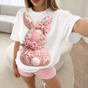 Puede incluir: Camiseta blanca con un diseño de conejo patchwork con patrones florales y cuadros rosas. El conejo tiene un lazo rosa, detalles de perlas y adornos de rosas. La modelo lleva shorts rosas.