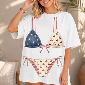 Könnte beinhalten: Weißes T-Shirt mit einem aufgedruckten Bikini-Design. Das Oberteil hat ein dreieckiges Denim-Design mit weißen Sternen und ein cremefarbenes Dreieck mit roten Kirschen. Die Unterseite hat eine cremefarbene Vorderseite mit roten Kirschen und einen rot-weiß gestreiften Rücken.