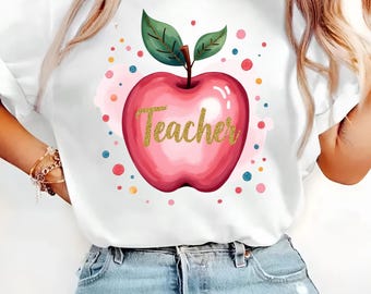 May include: White t-shirt featuring a large, pink apple graphic with the word "Teacher" in gold glitter lettering. The apple has green leaves and is surrounded by colorful polka dots, creating a playful design.