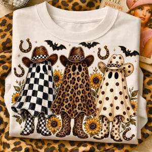 Könnte beinhalten: Ein weißes T-Shirt mit einer Grafik von drei Geistern, die Cowboystiefel und Hüte tragen. Die Geister haben Karomuster, Leopardenmuster und Punkte. Das Design enthält Sonnenblumen, Hufeisen und Fledermäuse im Hintergrund.
