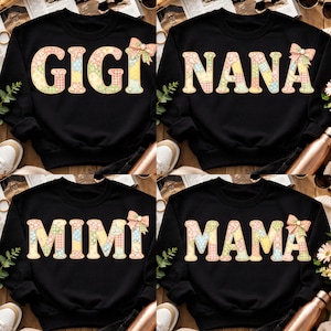 May include: Four black sweatshirts with the words "GIGI", "NANA", "MIMI", and "MAMA" in a patchwork design. Each word has a matching bow. The sweatshirts are displayed on a wooden surface with shoes and flowers.
