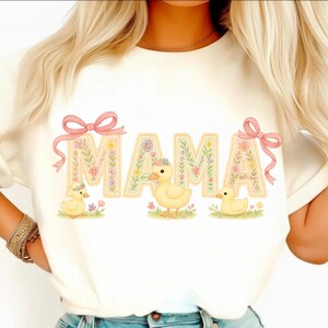 May include: Cream-coloured t-shirt with the word "MAMA" in a floral design, adorned with pink bows. Three yellow ducklings are beneath the letters, amidst grass and flowers. The top has a round neckline.