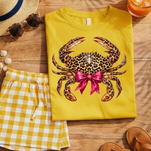 May include: Yellow sweatshirt with a leopard print crab design and a pink bow. The image also shows yellow and white checkered shorts, sunglasses, a straw hat, and sandals, suggesting a summer theme.