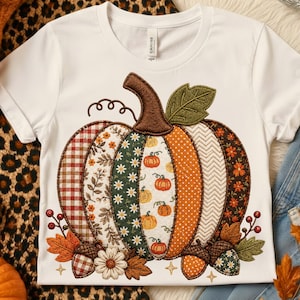 Könnte beinhalten: Ein weißes Kurzarm-T-Shirt mit einer großen, zentralen Kürbisgrafik, die aus verschiedenen gemusterten Stoffsegmenten wie Karos, Blumen und Punkten besteht. Das Design umfasst gestickte Eicheln, Blätter und kleine Blumen.