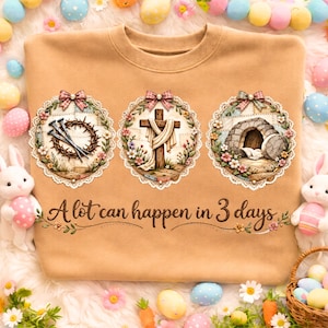 Puede incluir: Sudadera color beige con tres diseños circulares que representan símbolos religiosos: una corona de espinas y clavos, una cruz con un paño drapeado y una tumba vacía. Cada diseño tiene una corona floral y un lazo. El texto "A lot can happen in 3 days" está debajo.