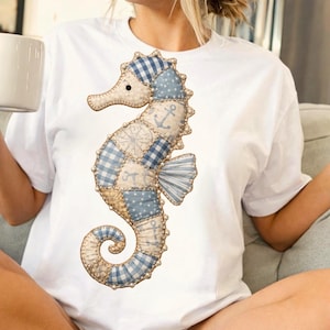 Pode incluir: Camiseta branca com um desenho de cavalo-marinho. O cavalo-marinho é feito de tecido patchwork em tons de azul e bege, com elementos náuticos como âncoras e bússolas. O cavalo-marinho é o elemento central do design.