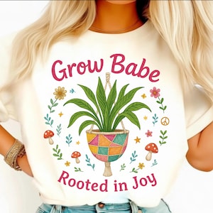 Puede incluir: Camiseta color crema con el texto "Grow Babe Rooted in Joy". El diseño presenta una planta colgante en una maceta de patchwork, rodeada de flores, setas y estrellas. Tema botánico.