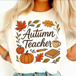 May include: A cream-coloured t-shirt with the words "Autumn Teacher" in a script font, encircled by autumn-themed illustrations. The design includes a pumpkin, apple, leaves, a coffee cup, a pencil, and a book.