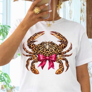 May include: White t-shirt featuring a detailed crab illustration. The crab has a leopard print shell and claws, with a bright pink bow. The shirt is a casual style, suitable for everyday wear.