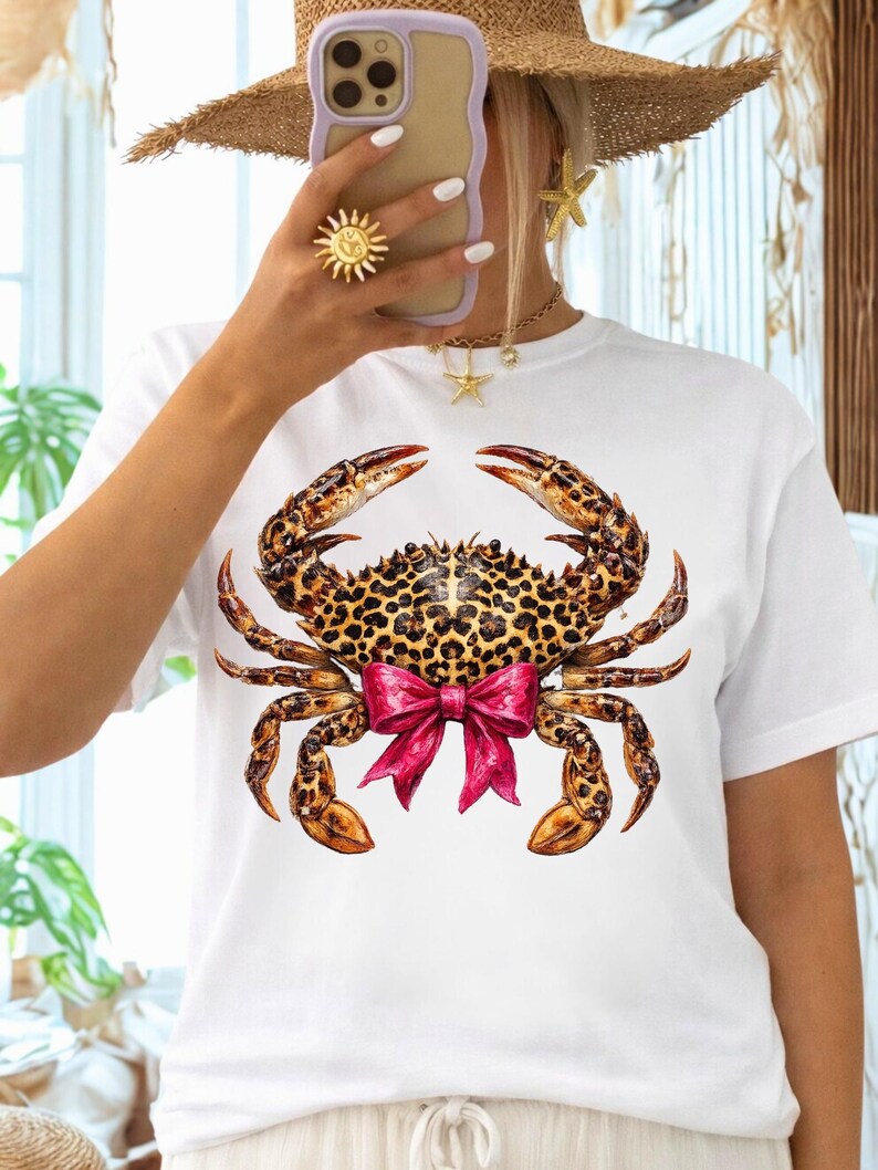 May include: White t-shirt featuring a detailed crab illustration. The crab has a leopard print shell and claws, with a bright pink bow. The shirt is a casual style, suitable for everyday wear.