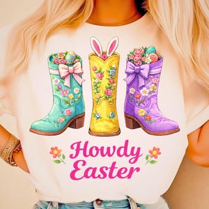 May include: A white t-shirt featuring three colorful cowboy boots with floral designs and Easter eggs. The boots are turquoise, yellow, and purple. The text "Howdy Easter" is in pink script below the boots.