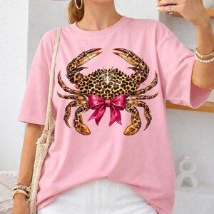 May include: A light pink t-shirt featuring a large crab graphic. The crab has a leopard print body, brown claws, and a pink bow. The shirt is a loose fit, with short sleeves.
