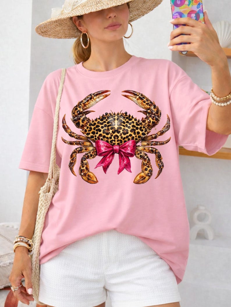 May include: A light pink t-shirt featuring a large crab graphic. The crab has a leopard print body, brown claws, and a pink bow. The shirt is a loose fit, with short sleeves.