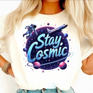 Op de afbeelding: Crèmekleurig T-shirt met een levendig kosmisch ontwerp. De afbeelding bevat een astronaut, een ruimteschip, planeten en de woorden "Stay Cosmic" in blauwe en witte letters. Het ontwerp is geplaatst tegen een donkerpaarse en blauwe ruimteachtergrond.