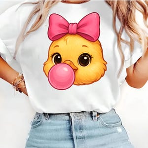 Könnte beinhalten: Weißes T-Shirt mit einem Cartoon-Küken-Design. Das Küken ist gelb und hat eine rosa Schleife und Kaugummi. Der Kaugummi hat eine leuchtend rosa Farbe. Das Design ist auf dem T-Shirt zentriert.