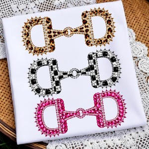 May include: A white t-shirt featuring three horizontal graphic prints of horse bit snaffles. The top bit has a leopard print, the middle bit is black and white checkered, and the bottom bit is solid hot pink. Each bit is outlined in rhinestones.