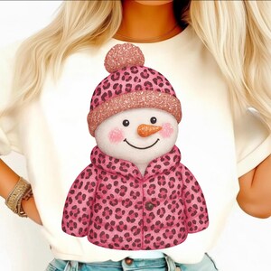May include: A white t-shirt featuring a whimsical snowman design. The snowman wears a pink leopard print hat and jacket with a matching pom-pom. The snowman has a carrot nose, rosy cheeks, and a smiling face.