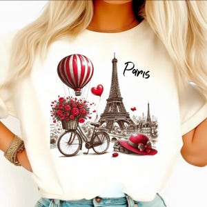 以下が含まれることがあります： パリをテーマにしたクリーム色のTシャツ。赤と白のストライプの熱気球、赤いバラのバスケットが付いた自転車、エッフェル塔、赤い帽子がデザインされています。「Paris」の文字は黒で書かれています。