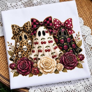May include: A white t-shirt featuring a graphic of three ghosts wearing large bows. The left ghost has a leopard print with skulls, the center is white with red cherries, and the right is black with pink skulls. Deep red and cream roses sit below.