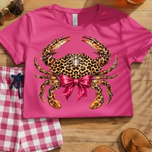 May include: A magenta t-shirt featuring a crab design with a leopard print body and a pink bow. The crab's claws and legs are detailed with brown and gold tones. A pair of pink and white checkered shorts and brown sandals are also visible.