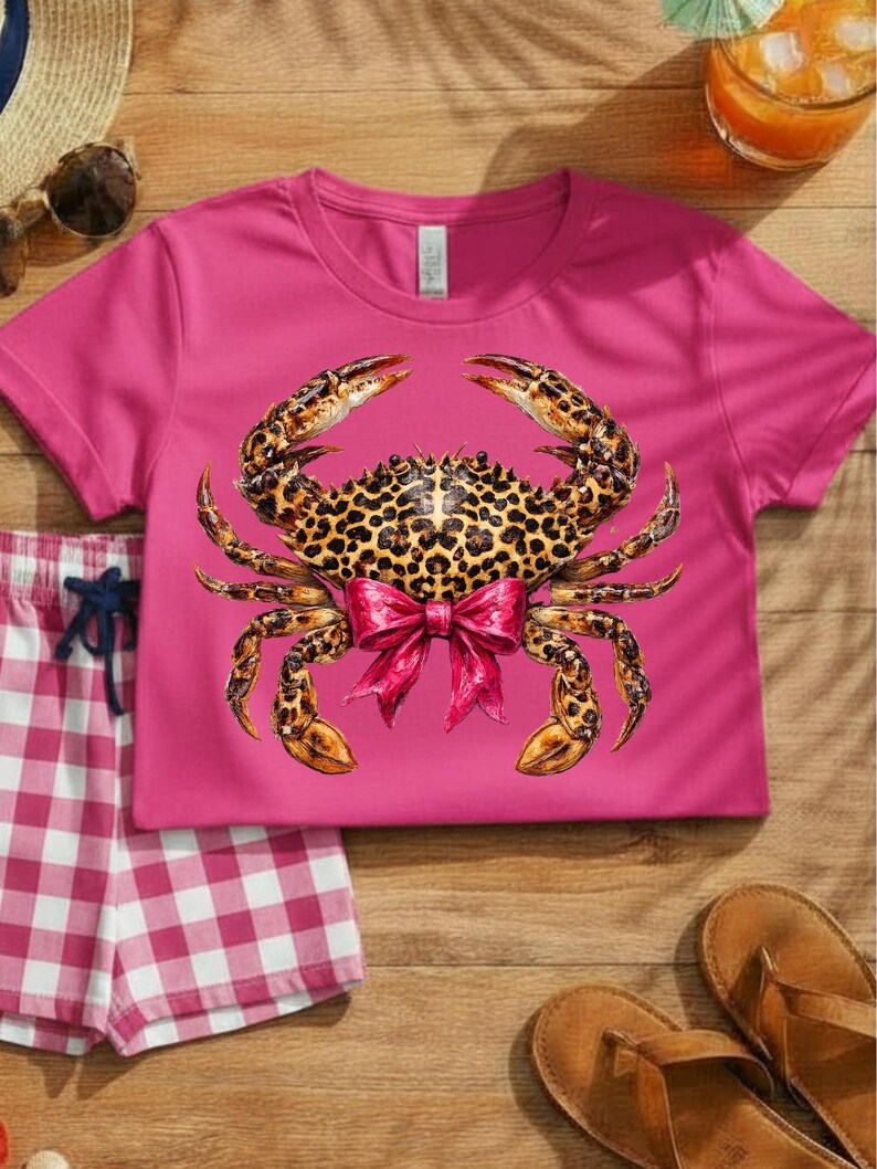 May include: A magenta t-shirt featuring a crab design with a leopard print body and a pink bow. The crab's claws and legs are detailed with brown and gold tones. A pair of pink and white checkered shorts and brown sandals are also visible.
