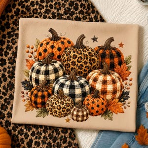 May include: A beige t-shirt featuring a graphic print of nine pumpkins in various patterns including orange and black buffalo plaid, leopard print, and polka dots, surrounded by autumn leaves, berries, and small brown stars.